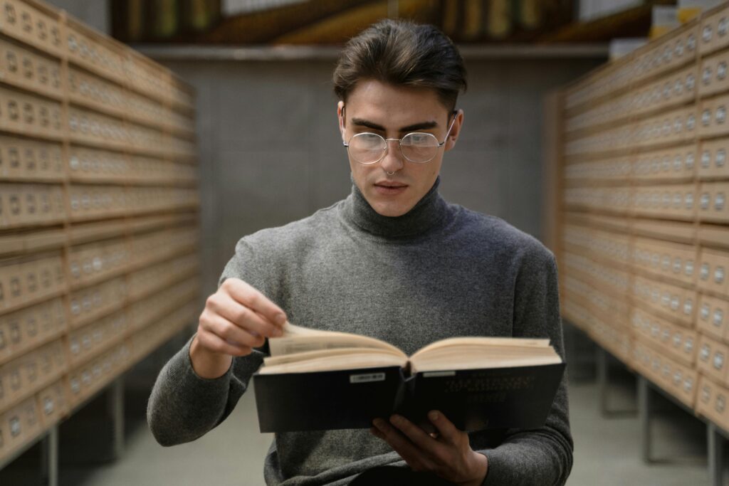 Young man in glasses reading a book in a library archive setting, focused on studying.