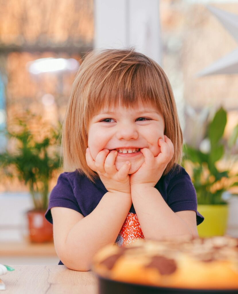 A happy young girl smiling indoors surrounded by plants and a pastry, with natural light streaming in.