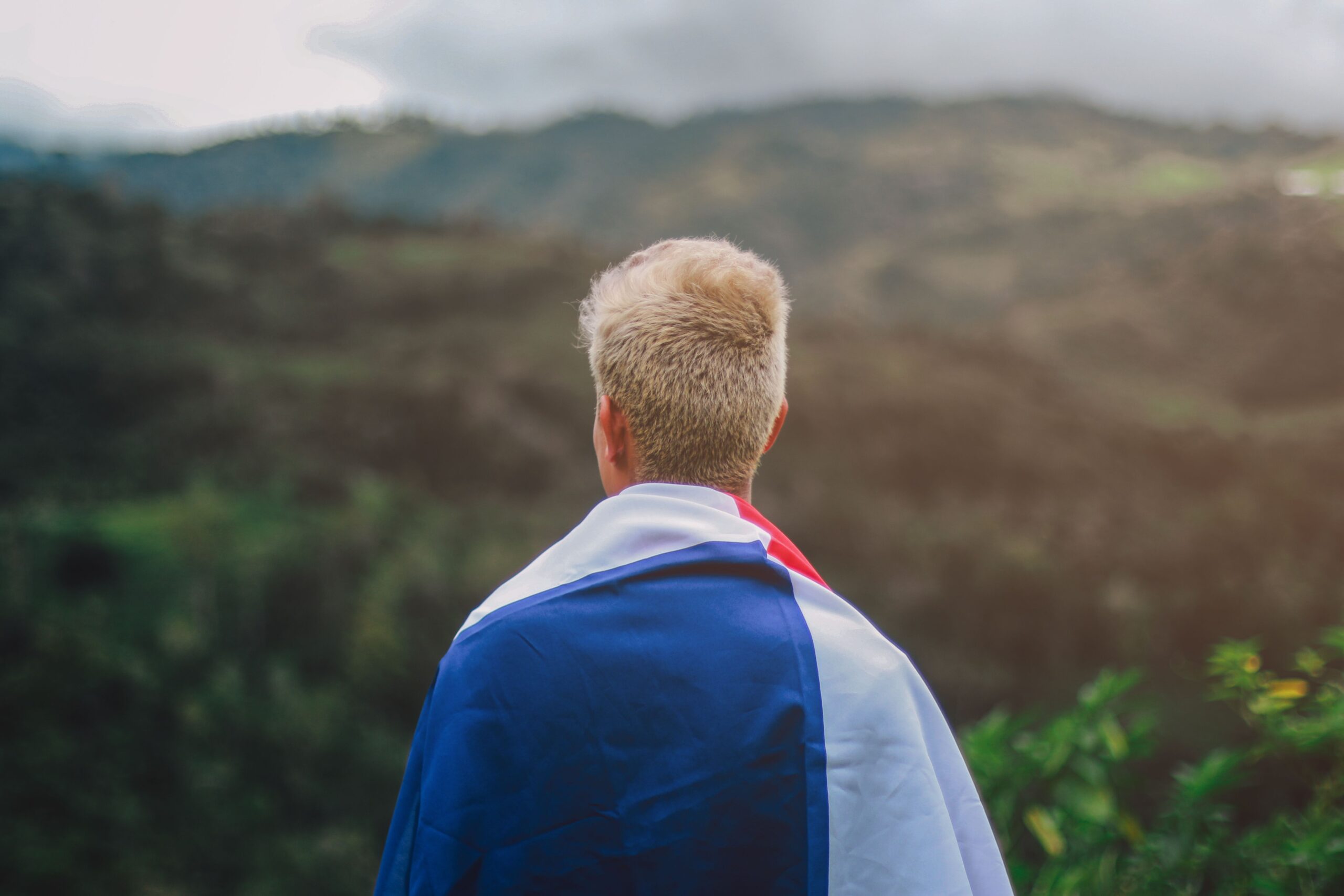 Back view of a person wrapped in a flag, gazing at a scenic mountainous landscape, symbolizing travel and exploration.