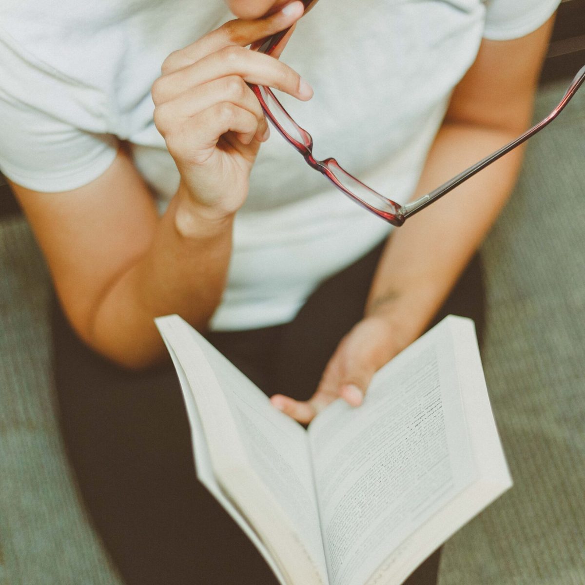 Young woman sitting indoors, reading a book while holding eyeglasses. A moment of focus and reflection.