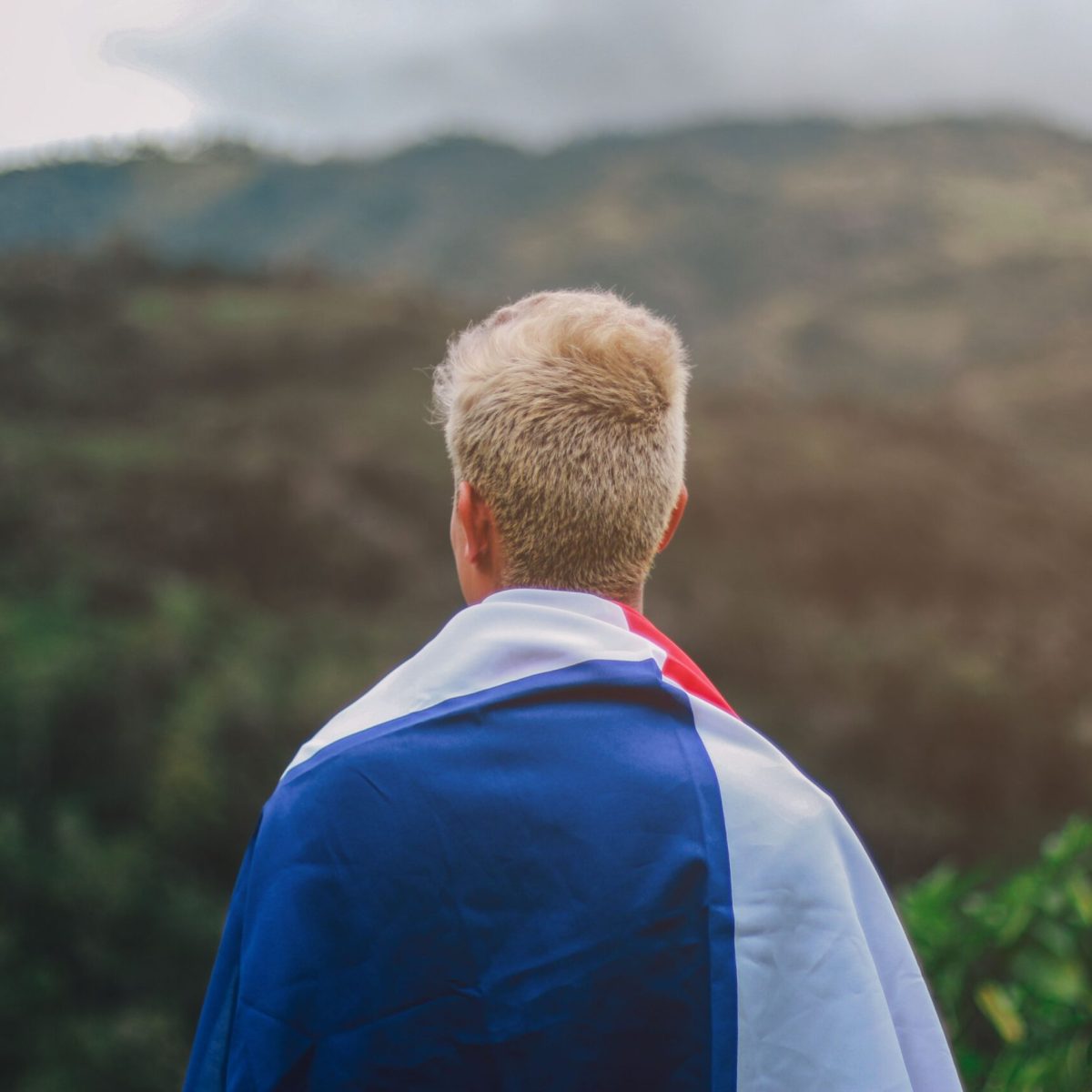 Back view of a person wrapped in a flag, gazing at a scenic mountainous landscape, symbolizing travel and exploration.